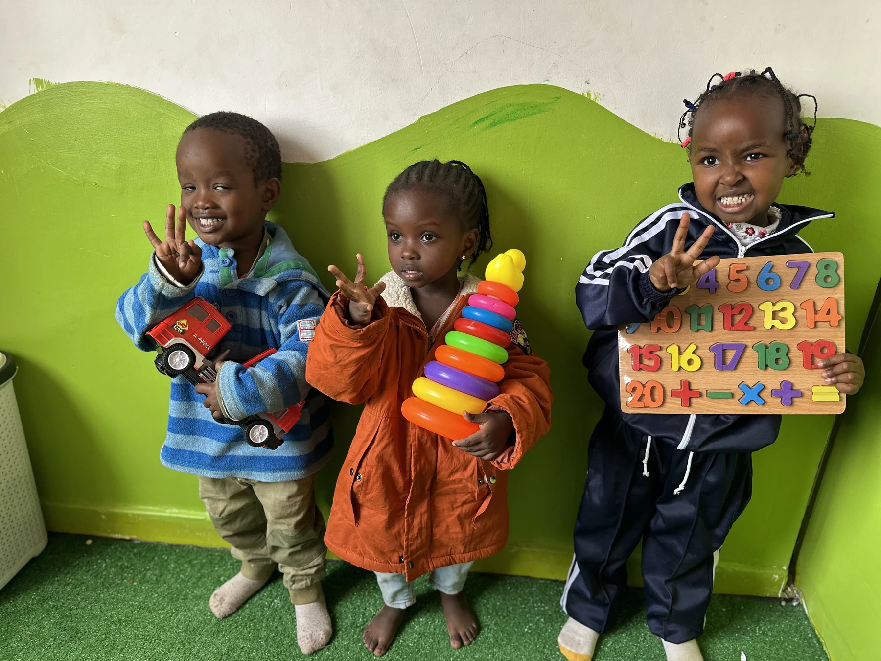 Three children showing a toy truck, stacking rings and a number board
